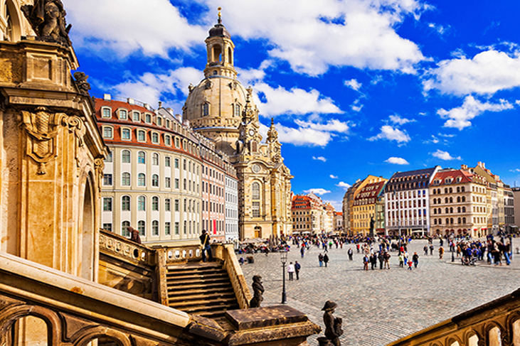 Barocke Frauenfigur auf einer steinernen Balustrade im Vordergrund, im Hintergrund die Frauenkirche Dresden mit umliegenden historischen Gebäuden und einem belebten Platz unter blauem Himmel mit weißen Wolken.