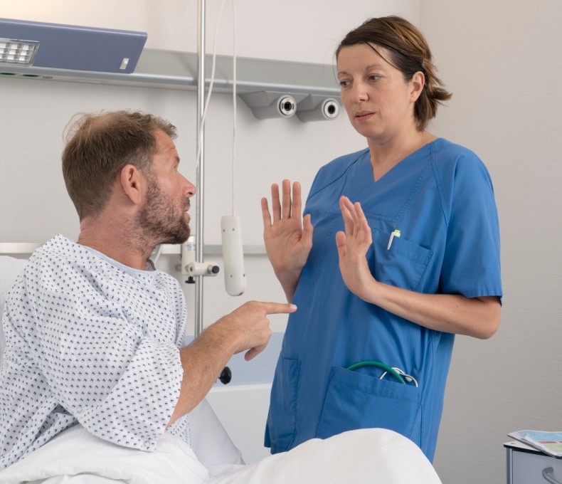 Eine Pflegerin weicht vor einem Patienten zurück, der mit dem Finger auf sie zeigt. Sie hebt abwehrend die Hände. Die Pflegerin trägt einen blauen Kittel. Der Patient ein Krankenhausnachthemd. 