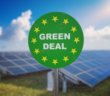 Auf einem grünen runden Schild steht: Green Deal; im Hintergrund sind Solarpanele zu sehen