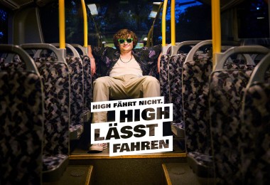 Das Bild zeigt einen jungen Mann mit lockigem Haar und grüner Sonnenbrille, der lässig auf einem Sitz in einem fast leeren Bus sitzt. Seine Arme sind entspannt über die Rückenlehnen der Sitze gelegt. Er trägt eine helle Hose, ein weißes T-Shirt und ein dunkles Hemd mit Muster. Im Vordergrund steht der Text „HIGH FÄHRT NICHT. HIGH LÄSST FAHREN.“ auf weißen, leicht schräg angeordneten Blöcken. Die Innenausstattung des Busses ist mit gemusterten Polstern gestaltet, und das Bild vermittelt eine entspannte, abendliche Atmosphäre.