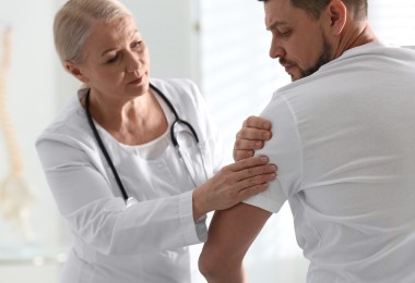 Ärztin in weißem Kittel untersucht die Schulter eines sitzenden Mannes. Sie tastet den Oberarm ab, während der Mann schmerzhaft den Kopf abwendet. Beide tragen weiße Kleidung. Im Hintergrund ist unscharf ein Modell einer Wirbelsäule zu erkennen.