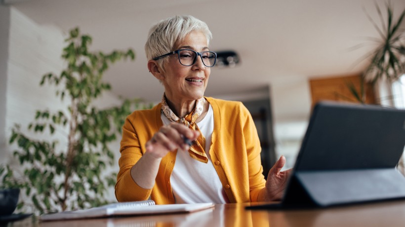 Ältere Frau mit kurzem grauen Haar, Brille und gelber Strickjacke sitzt an einem Tisch und schaut lächelnd auf ein Tablet. In der Hand hält sie einen Stift, daneben liegt ein Notizbuch. Die Umgebung wirkt wohnlich, im Hintergrund sind Pflanzen zu sehen.