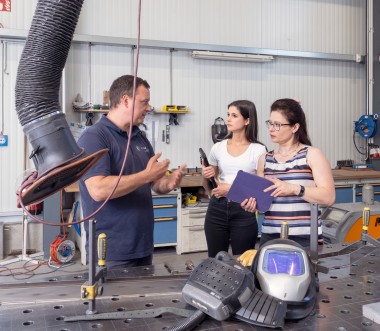 In einer Werkstatt mit metallverarbeitender Ausstattung stehen ein Mann und zwei Frauen im Gespräch. Der Mann trägt ein dunkelblaues Poloshirt und erklärt gestikulierend etwas. Die eine Frau hält ein Klemmbrett, die andere ein Werkzeug. Im Vordergrund liegt ein Schweißhelm auf einem großen Metallarbeitstisch, an dem Absaugvorrichtungen und weitere Werkzeuge angebracht sind. Im Hintergrund sind Werkbänke, Gasflaschen und weitere Arbeitsmittel sichtbar. 