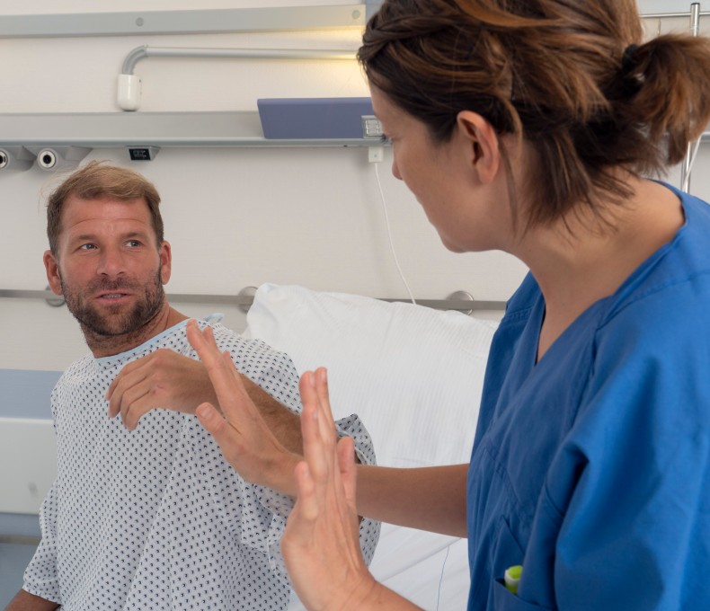 Ein Mann in Krankenhauskleidung sitzt aufrecht im Bett und schaut eine Pflegekraft an, die ihm gegenübersitzt und mit erhobenen Händen eine abwehrende Geste macht. Die Situation wirkt angespannt.