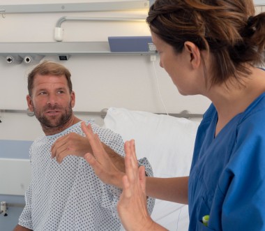 Ein Mann in Krankenhauskleidung sitzt aufrecht im Bett und schaut eine Pflegekraft an, die ihm gegenübersitzt und mit erhobenen Händen eine abwehrende Geste macht. Die Situation wirkt angespannt.