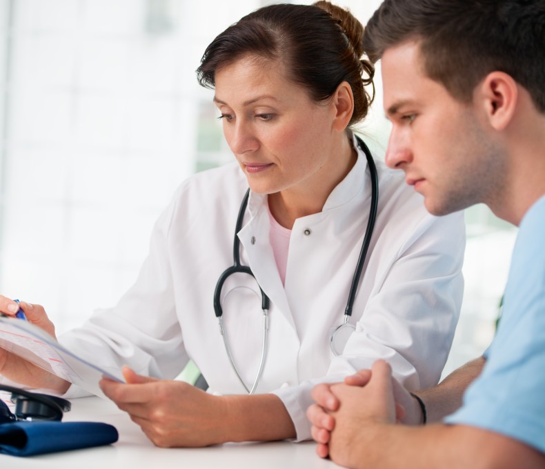 Eine Ärztin in weißem Kittel mit Stethoskop um den Hals sitzt an einem Tisch und bespricht ein Formular mit einem jungen Mann. Beide blicken konzentriert auf das Dokument. Auf dem Tisch liegen ein Blutdruckmessgerät und ein Kugelschreiber.