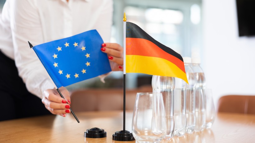 Eine Person im weißen Hemd mit rot lackierten Fingernägeln stellt zwei kleine Tischflaggen auf einem Konferenztisch auf: eine blaue Flagge der Europäischen Union mit gelben Sternen und eine schwarz-rot-goldene Flagge Deutschlands. Daneben stehen mehrere Wasserflaschen und leere Gläser.