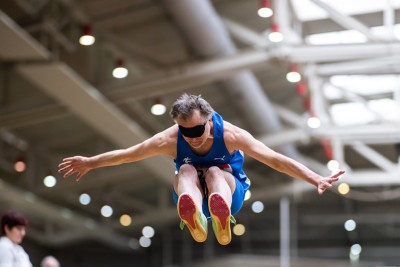 Ein Weitspringer kommt direkt auf uns zugesprungen. Beine angewinkelt, Oberkörper nach vorne gelehnt, Arme nach links und rechts ausgestreckt. der Springer ist älter und trägt eine Augenbinde.